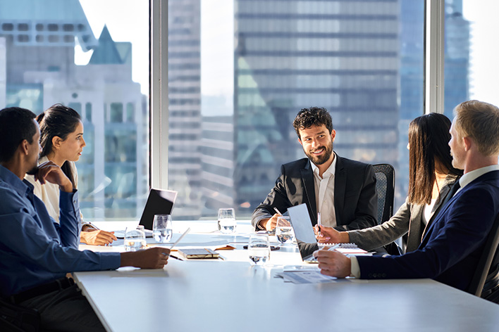 Photo of 5 businessmen and businesswomen around a desk discussing business in a conference room