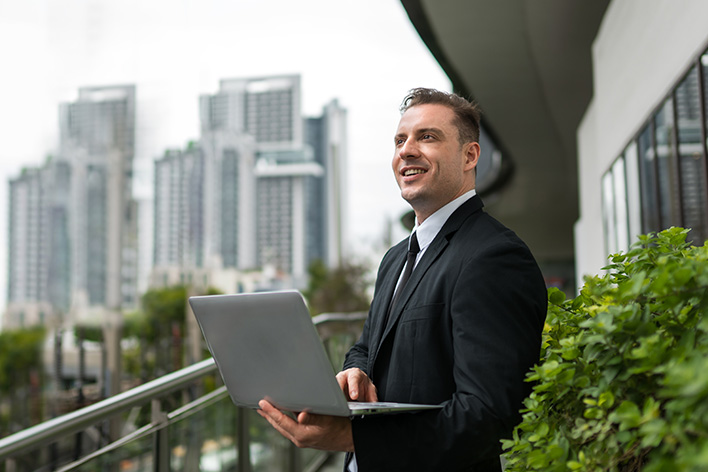 Photo of a businessman in a suit holding his laptop on an outdoor balcony